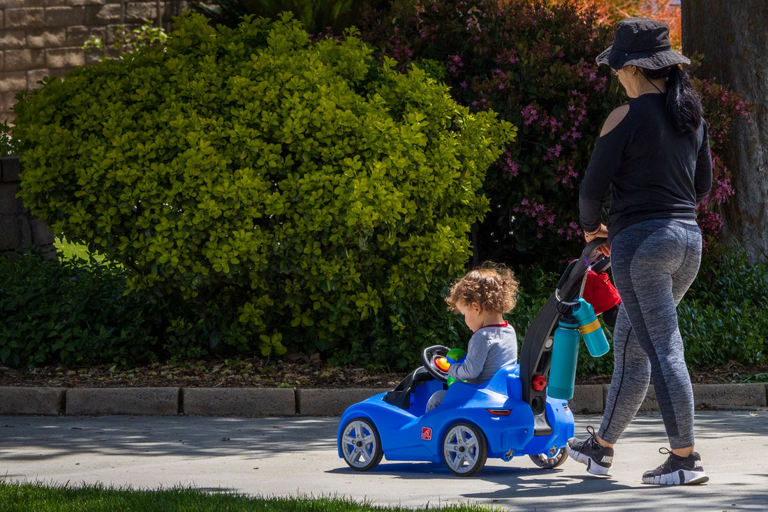 Parent walking alongside a toddler riding a toy car outdoors