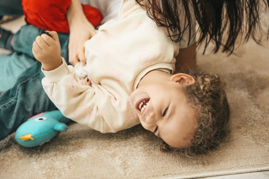Toddler laughing on the floor with a plush toy and a parent nearby